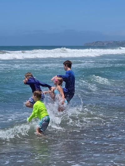 This is a full-frame, eye-level, outdoor, vertical, natural light photograph of four children playing in the ocean waves at a beach. The two older children, a boy and a girl, are on the right side of the frame. The boy, wearing a blue long-sleeved rash guard, is reaching out and splashing. The girl, wearing a blue bikini and pink goggles, is in the center, also splashing. To the left, a younger boy in a blue long-sleeved rash guard and patterned swim trunks is jumping or lunging forward, with goggles on. In the foreground on the left, a young boy in a bright yellow long-sleeved rash guard and patterned swim trunks is visible, also playing in the waves. The ocean is a deep blue-green with foamy white waves breaking. In the background, the sky is a clear blue with some hazy clouds. A distant cliff face, possibly representing the coastline of San Clemente, California, is visible on the horizon. The overall scene captures a moment of joyful activity and play in the ocean.