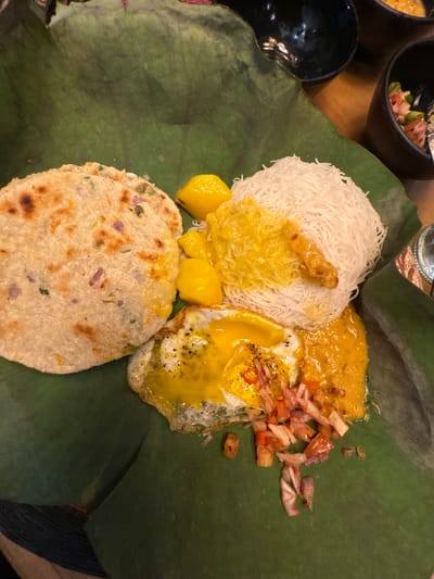 The image shows a Sri Lankan meal served on a green leaf. The meal includes string hoppers (a type of noodle made from rice flour), a curry or stew, fried egg, and a flatbread. The components are arranged neatly on the leaf. This type of meal is commonly served in Kandy, Sri Lanka. The surrounding area shows bowls and what appear to be parts of a wooden table.