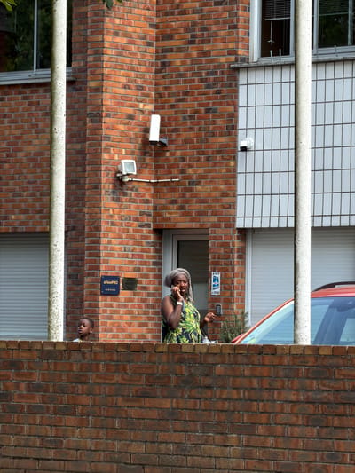 The image captures a vertical view of an exterior wall, predominantly constructed from reddish-brown bricks. A thick, light-colored pipe or railing runs horizontally across the foreground, obscuring parts of the scene. The upper section of the building features a section with white square tiles.
Two individuals are visible. In the mid-ground, a person with dark skin is leaning against the brick wall, wearing a patterned garment with green and yellow foliage designs. They are touching their face and appear to be engaged in conversation or observation. Further down, near the bottom right, another person, also with dark skin and wearing a yellow collar shirt, peeks out from behind the brick wall. Their head is tilted downwards, suggesting they are looking at something out of frame or resting.
Several objects are mounted on the brick wall: two white security cameras, one a cylindrical shape, the other a more boxy design with a visible lens. Below the cameras, a blue and white sign reads "AFMPRO" with a phone number "06 98 72 39 54" below it. Another smaller, darker plaque is positioned above the AFMPRO sign. A glimpse of a red vehicle is visible in the upper right portion of the image, near a window.
The scene appears to be outdoors, likely in an urban or residential setting given the building's construction and the presence of security cameras. The lighting suggests it is daytime, with shadows cast by the architecture. The overall atmosphere is calm and uneventful, with the individuals seeming to be momentarily paused.