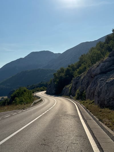 A winding asphalt road cuts through a mountainous landscape in Bukovica, Montenegro. The road, marked with a solid white line on the right and a dashed white line on the left, curves through the scene, disappearing into the distance. On the right side of the road, a steep, rocky mountainside covered in green trees rises sharply. On the left, gentler, tree-covered slopes recede into the background, with layered mountains visible against a clear, bright blue sky. The sun is high, casting a bright glare in the upper right portion of the image, and illuminates the road surface, highlighting its texture and the white markings. There are no people or vehicles visible in the image. The environment is natural and rugged, characterized by the mountainous terrain and dense vegetation.