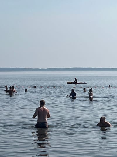 In the city of Surakovo, Russia, a group of individuals are engaged in recreational activities on a body of water, likely a lake or large river, under a clear, overcast sky. The water is calm, with subtle ripples reflecting the muted light. In the foreground, a shirtless man stands facing away from the camera, his back to the viewer, with his lower body submerged in the water. Further out, a collection of people are scattered across the water. Some are standing or wading, while others appear to be swimming or relaxing. One individual is seated on a paddleboard in the mid-ground, facing the shore. A line of buoys or markers is visible in the water, potentially delineating a swimming area or a course. The distant background shows a faint, tree-lined horizon under the pale sky, suggesting a natural or semi-natural environment. The overall atmosphere is one of leisure and outdoor recreation.