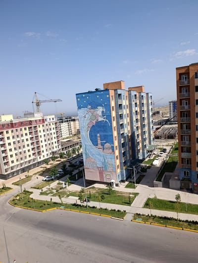 This is a high-angle, daylight exterior shot of a newly developing urban area in the city of Yangi Andijan, Uzbekistan. In the foreground, a wide asphalt road curves to the left, bordered by a yellow curb and green landscaping with small trees and shrubs. Several vehicles are visible on the road and in parking areas, including white sedans and a white van.

Dominating the midground are several modern, multi-story apartment buildings. The building on the far right is orange and white. Next to it is a cluster of three taller, narrow buildings, painted in alternating bands of blue and orange, with balconies. The leftmost of these blue and orange buildings features a large, vibrant mural covering its entire side. The mural depicts a historical or mythological scene with a rider on a horse within a crescent moon, a mosque with a minaret, and a domed structure, all set against a starry blue sky.

Further in the background, more apartment buildings are under construction, with cranes and scaffolding visible. The overall impression is one of rapid urban development. The sky is clear and blue with a few wispy clouds. The environment appears to be a residential district with amenities such as walkways and green spaces.