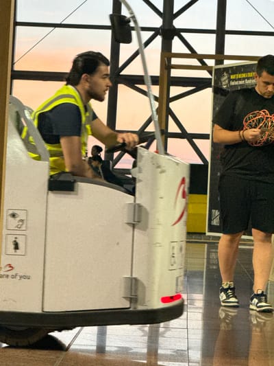 The image shows a man in a neon yellow vest driving a small vehicle, likely in an airport terminal. The man has dark hair and a beard and is wearing a dark t-shirt under the vest. The vehicle is white with some red markings and appears to be a cargo or baggage handler. In the background, another man in a black t-shirt and shorts is standing and holding a decorative object made of wire. Behind him, a poster with text is visible, including "WANT TO BECOME A TRUE FL... Find out more: flan...". The environment appears to be indoors with large windows showing an orange sky, suggesting either sunrise or sunset. The flooring is reflective, indicating a polished surface. This scene is likely at Brussels Airport (BRU) in Zaventem, Belgium.