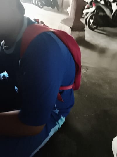 The image shows a person from the back, wearing a blue shirt with red accents. The person appears to be a child, possibly kneeling or bending over. The environment seems to be an outdoor urban setting, with what appears to be a concrete pillar and a blurred background of motorcycles. The lighting is dim, suggesting it might be evening or an indoor dimly lit area. The image is grainy and not very clear. There is no visible indication of the location being Katrungan, Indonesia.