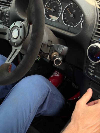 The image shows a close-up view from the driver's perspective inside a vehicle. The focus is on the steering wheel, dashboard, and the driver's legs. The steering wheel is wrapped in dark material with red stitching visible on the bottom section. The dashboard features various gauges, including a speedometer and a tachometer, and several buttons and knobs for climate control and other functions. A car key is inserted into the ignition. The driver is wearing light blue pants. A red racing shoe with black accents is visible on the driver's right foot, resting on or near the pedals. The overall environment suggests the interior of a car, possibly a performance-oriented one given the steering wheel and racing shoe. There is no visual information to determine the specific location within Навадворскі сельскі Савет, Belarus.