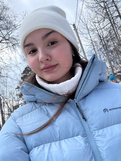 The image displays an upward-angled selfie of a young female in a winter outdoor setting. The primary subject, centrally positioned, has fair skin, dark hair, and wears a white knit beanie, a white ribbed turtleneck, and a light blue quilted puffer jacket featuring a visible "OUTVENTURE" logo on the right chest. A small, clear stone earring is present in her left ear. Her expression is a slight smile directed towards the camera. In the upper right background, a second individual is partially visible, wearing a blue and white patterned knit beanie. The environment consists of numerous leafless deciduous trees, suggesting a forest or park. An overcast, light grey sky is visible. A grey utility pole with wires stands in the left background. The image provides no specific visual identifiers for the city of Nikola, Russia.