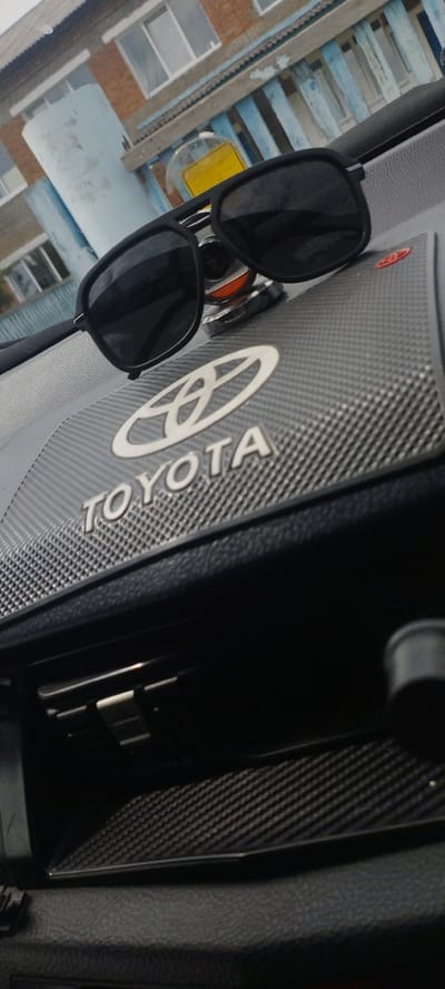 The image shows a pair of black sunglasses resting on a black textured dashboard with a silver Toyota logo. The dashboard is inside a vehicle. In the background, a light blue cylindrical object and a building with windows are visible through the windshield, suggesting an outdoor setting. The building has a textured facade and a blue railing. The environment appears to be urban or suburban, likely in a location with moderate weather conditions. The lighting suggests it is daytime. The car's interior, specifically the dashboard area, is the primary focus. No people are visible in the image. The location is identified as Pokosnoye, Russia.