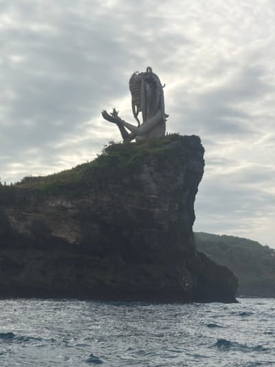 A large, grey-toned statue depicting a humanoid figure with long, intricate dreadlock-like hair and ornate headwear is positioned atop a rugged, dark grey cliff overlooking a body of water. The figure is shown from approximately the waist up, with both arms raised and hands open, palms facing upwards, possibly holding a small, indistinct object between them. The cliff face is steep and rocky, with sparse green vegetation on its upper surface. Several small human figures are visible near the base of the statue, providing scale, and a faint fence or railing runs along the cliff edge where they stand. The water below is dark blue-grey with visible ripples and small waves. In the background, under an overcast sky with grey and white clouds, another green-covered landmass extends. This scene is located in Sakti, Indonesia.