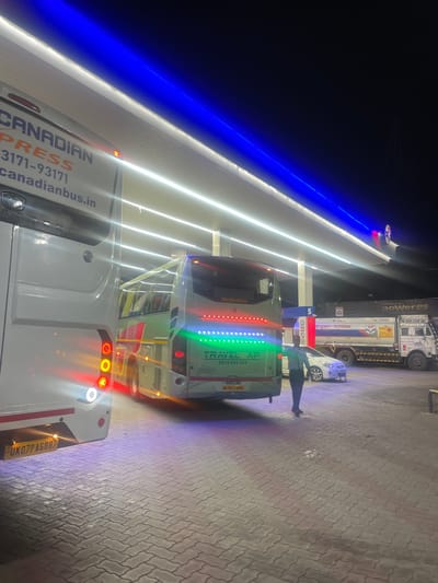 At night in Kajheri, India, a gas station is illuminated by bright lights. A white bus labeled "CANADIAN EXPRESS" is parked on the left, its rear lights reflecting on the wet pavement. To its right, another green bus with "TRAVEL AF" printed on it is also parked. A man in a light blue shirt and dark pants stands between the buses, facing towards the right. Further to the right, a white car and a large tanker truck with "Hindustan Petroleum" branding are visible. The ground is paved with interlocking bricks, and the sky is dark, with blue and white lights from the gas station canopy.