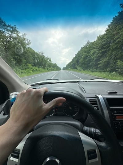 The image shows a first-person perspective from inside a car, with the driver's hands on the steering wheel. The car is on a road, with lush green trees lining both sides of the road. The sky is visible through the windshield, with a mix of blue sky and clouds, suggesting it's daytime. A can with "L-CARNITINE VIT" is visible near the bottom of the frame. The dashboard and steering wheel of the car are also visible. The scene appears to be a road trip through a rural or natural area. The weather seems to be fair, with a blend of sunshine and clouds. The image is oriented sideways.