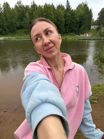 A woman is taking a selfie by a body of water. She is wearing a pink and blue hoodie with a Hello Kitty graphic on the front. The water is calm with ripples indicating rain. Behind her is a wooded area and a few small buildings. The location is identified as Novaya Vozhoika, Russia.