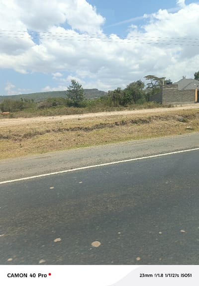 The image depicts a static outdoor scene in Gilgil ward, Kenya, characterized by a mix of infrastructure and natural environment under a partly cloudy sky.

In the immediate foreground, a dark asphalt road spans the lower third of the frame, showing signs of dirt and light-colored spatters. A faded white line delineates the right edge of the visible road. Above the road, a dirt shoulder or unpaved track extends horizontally across the midground, covered predominantly with dry, light-brown grass and sparse, low-lying green vegetation.

In the background, on the left side, undulating hills or mountains covered with scrub brush are visible, rising against the horizon. A light-brown animal, possibly a cow, is partially observable standing on the dirt track in this area. Towards the right side of the background, a partially constructed or recently completed building is present. It features grey concrete block walls and a pitched roof with grey tiles. A black water tank is situated on the roof. Various trees and bushes are scattered throughout the background, particularly around the building and along the base of the hills. Overhead, several thin, dark horizontal lines, consistent with power or telephone lines, traverse the sky. The sky itself is clear blue with numerous white cumulus clouds. No human figures or active interactions are visible in the scene.