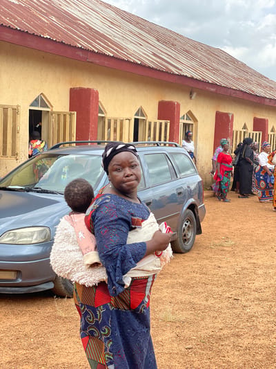 The image captures an outdoor scene in the city of Kassa, Nigeria, with a building that appears to be a community hall or church serving as the backdrop. The building has a corrugated metal roof, faded yellow walls, and red accent pillars. Several windows with wooden shutters line the facade, some open revealing figures inside. In the foreground, a woman is carrying a baby on her back, wrapped in a patterned cloth and a fluffy cream-colored carrier. She is wearing a dark blue dress with intricate patterns and a black and white headscarf. Beside her is a gray station wagon, its doors and windows reflecting the environment. Behind the woman and the car, a group of other women are gathered, dressed in colorful African attire, suggesting a communal event or gathering. The ground is covered in reddish-brown dirt, indicative of a rural or semi-urban Nigerian setting. The sky above is overcast with clouds.