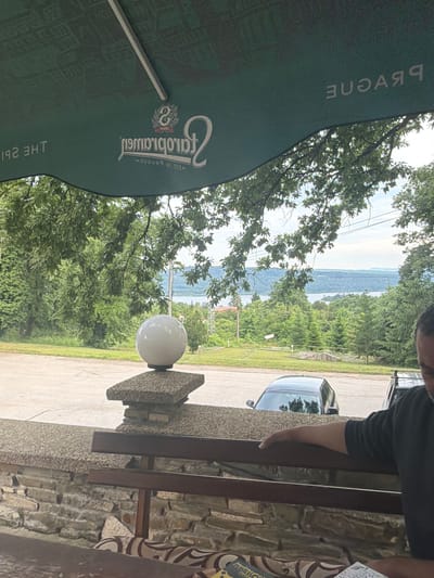The image depicts an outdoor scene, likely from a restaurant or cafe patio, offering a panoramic view of a large body of water, possibly a river or lake, surrounded by densely forested hills. The sky above is bright and appears somewhat hazy.

In the foreground on the left, a large dark green outdoor umbrella shades part of the area. Text is visible on the umbrella, printed in reverse: "PRAQNE" (which would be "PRAGUE" when read correctly), and a prominent logo featuring the word "Gambrinus" along with the year "1869." Other partially visible, reversed text includes "THE SPI..." and "EANRAB IH ТЭ2" (likely "THE... BARNE" or similar).

To the right, a person wearing a dark top is partially visible, with their arm resting on a stone railing or wall. Below the railing, a dark-colored car is parked on a paved surface. A white spherical outdoor lamp is mounted on a square stone base, positioned in the mid-ground. The architecture includes a rough stone wall or facade on the right.

The scene conveys a relaxed, daytime atmosphere, suggesting a pleasant spot to enjoy the scenic landscape of water and green hills. The presence of the Gambrinus beer logo on the umbrella indicates it's a commercial establishment. The setting aligns with a natural, possibly rural, landscape like that found near Sushina, Bulgaria.