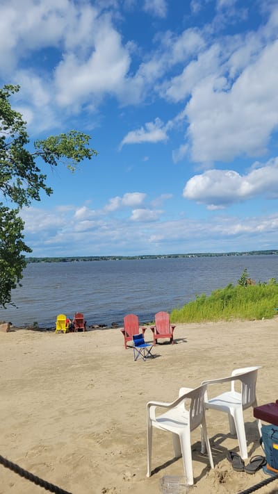 The image depicts a sandy beach area with a large body of water in the background, likely Lake Saint-Louis in Pointe-des-Cascades, Canada. The sky is blue with scattered white clouds. Several Adirondack chairs are present on the beach: two red ones are positioned in the foreground, with a small blue folding chair between them. Further back, closer to the water's edge, are two more Adirondack chairs, one yellow and one red, with two people seated in the red chair. A green tree branch with leaves extends into the upper left corner of the frame. The water appears slightly choppy. In the lower right portion of the image, two white plastic chairs face away from the viewer, with a pair of flip-flops on the sand in front of them and a cooler next to them. The sand is textured with footprints.