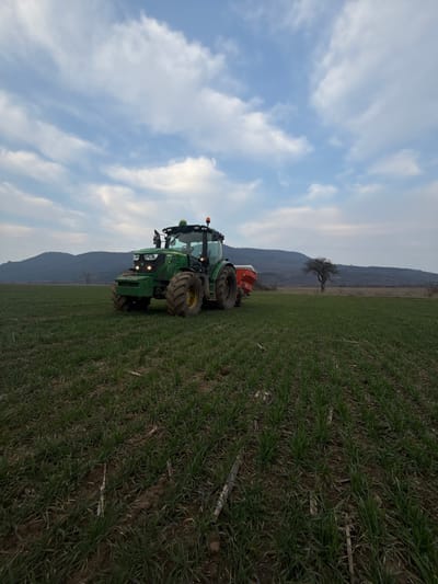 The image displays an agricultural field located in Gagovo, Bulgaria, under a partly cloudy sky. A green John Deere tractor, equipped with large, lugged tires, a driver's cab, and two amber warning lights on its roof, is positioned in the mid-ground, angled slightly to the left. Its front headlights are illuminated. A red agricultural implement, consistent with a fertilizer spreader or seeder, featuring a white top, is attached to the rear of the tractor. The foreground and mid-ground of the field are covered in orderly rows of young, green crops, with some dried plant matter visible between the rows. To the right in the mid-ground, a solitary, leafless deciduous tree with dark branches stands. A range of dark-hued hills or mountains forms the distant background horizon. No individuals are present in the scene.