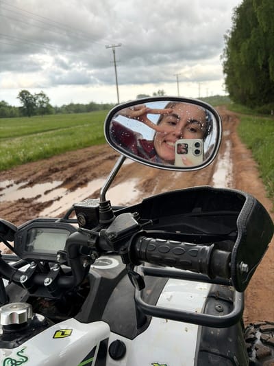 The image depicts a view from the front of an all-terrain vehicle (ATV). The focus is on the ATV's handlebars, gauges, and a side mirror. The mirror reflects a young woman taking a selfie, holding up a peace sign. Water droplets are visible on the mirror's surface. Behind the ATV, a muddy dirt road stretches into the distance, flanked by green fields and trees. Utility poles are also visible in the background. There is no indication of the location.
