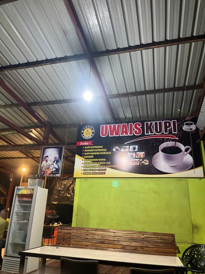 The image displays a sign for "UWAIS KUPI" with a coffee cup graphic and a list of available drinks. The menu includes Kupi Expresso, Sanger Expresso, Kupi Sareng Robusta, Sanger, Boh Manok Weng, various juices and instant coffees, Cappuccino Cincau, and Lemon Jahe Madu. The sign hangs from the ceiling of what appears to be a shop or cafe.