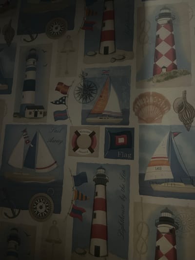 The image is a photograph of wallpaper with a nautical theme. The wallpaper features various illustrations of lighthouses, sailboats, anchors, compasses, flags, shells, and a life preserver. The colors are predominantly blues, whites, and reds, with some beige and brown accents. The overall impression is one of a decorative pattern. There are no people visible in the image, and the setting appears to be indoors, as suggested by the flat, wallpapered surface. There is no information in the image to suggest a specific location in Kalijaten, Indonesia, or any other city. The photograph is taken from a slightly elevated angle, with the lighting appearing somewhat dim, creating shadows and highlighting some areas more than others. The image has a slightly grainy texture.