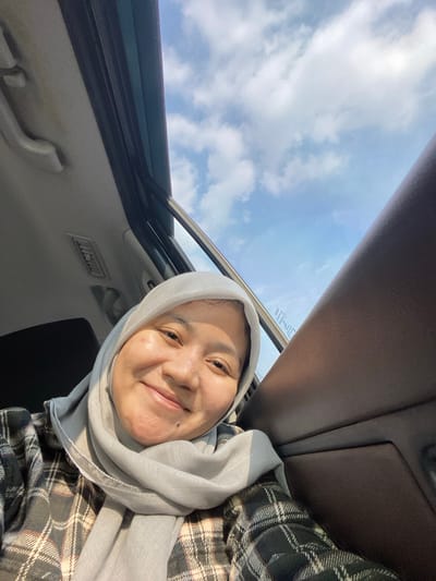 A medium shot, selfie taken from inside a car, shows a smiling woman wearing a light grey hijab and a plaid shirt. She is seated on the right side of the car, with her face turned towards the camera and sunlight illuminating her features. The car's interior is visible, including the roof lining, air vent, and a dark brown car door panel. The sunroof is open, revealing a bright blue sky with scattered white clouds. The image appears to be taken during the daytime. The specific location in Mandar Sari, Indonesia, is not discernible from the image itself, but the setting suggests a journey within or near a populated area, likely a city or town.