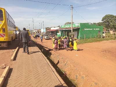 This image captures a sunny daytime scene in Shirungu-Mugai ward, Kenya, depicting a semi-urban roadside environment. On the far left, multiple yellow buses are parked or stationary; the closest bus displays "ONE" on its lower rear panel. Adjacent to the buses, a red-brown brick-paved sidewalk with a herringbone pattern extends into the foreground. A male subject, dressed in a dark suit, walks away from the viewer on this sidewalk.
An open, shallow drainage ditch separates the paved sidewalk from a wide, unpaved, red-brown dirt road that occupies the center and right foreground. On this dirt road, a group of approximately 10-15 individuals, primarily women, are congregated. Many in this group wear matching yellow attire, including hats or head coverings, while others wear pink or patterned dresses. Additional individuals, also wearing yellow, are visible further down the dirt road.
Across the dirt road, a line of single-story commercial buildings is visible. The most prominent structure is bright green with "Safaricom m-pesa" displayed in white lettering, indicating a mobile money service point. Utility poles supporting overhead power or communication lines are distributed throughout the scene. In the mid-ground on the dirt road, several vehicles, including a white van, are present. Sparse trees and vegetation are observed beyond the commercial buildings on the right and in the background. The sky above is clear blue.