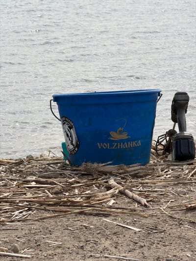 The image depicts a blue Volzhanka fishing bucket and fishing equipment on a riverbank near Krasnyi Bor, Russia. The bucket is positioned adjacent to the water. The ground is covered with dry reeds and earth.