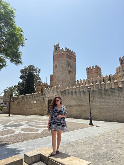 A woman in a black and white dress stands on patterned pavement next to a tall, stone castle or fortress with crenellations. The building has a weathered appearance. A lamp post is situated nearby. Trees are visible in the lower left of the frame. The sky is clear and blue. The perspective is slightly tilted.