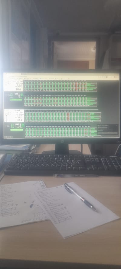 This media file depicts a close-up, slightly angled shot of a computer monitor displaying technical data, likely in an industrial or control room setting. The screen shows a grid of green and red circles, representing status indicators, alongside numerical data presented in rows and columns. There are labels indicating "ТП-1" and "ТП-2," which could refer to technical parameters or substations. Textual elements on the screen include values for "P" (power), "Q" (reactive power), "U" (voltage), and frequencies in Hertz (Гц) and Kilovolt-Ampere reactive (кВар). Some of the numerical values are large, such as 1245.6 kW and -1324.7 kW.
To the right of the monitor, a black keyboard is visible, suggesting a user interaction point. A pen rests on a piece of paper with handwritten notes, further indicating an active work environment. The paper contains a mix of numbers and illegible script, possibly related to the data displayed on the screen or a separate task.
The scene appears to be indoors, with the monitor and keyboard placed on a desk. The lighting is somewhat dim, with reflections visible on the monitor screen, hinting at the presence of other light sources in the room. The overall impression is of a functional workspace focused on monitoring and managing technical systems. The specific location context of Ushtobe, Kazakhstan, combined with the technical data on the screen, suggests this could be a control room at a power substation or industrial facility in that region. There are no people visible in the image.