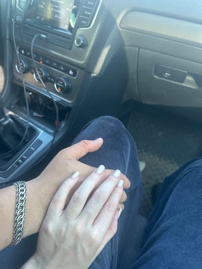 The image shows a close-up view of two people holding hands inside a car. The hands are resting on dark blue clothing. One hand wears a silver bracelet with a chain-link design. The background shows the interior of a car, including the dashboard with a radio display and climate controls, and a gear shift. The car appears to be parked, as evidenced by the static position of the elements within. The lighting suggests it is daytime. The scene is devoid of any specific landmarks that would place it in Vasilyevo, Russia.