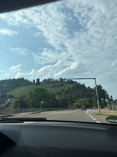 This is a landscape image, taken from inside a vehicle, showing a view of Gernsbach, Germany. The scene is dominated by a lush green hillside covered in vineyards, stretching upwards and dotted with trees. On top of the hill, there appears to be a historical building or castle. The sky is a bright blue, filled with scattered white clouds.
To the right of the hillside, a paved road runs horizontally across the image. There are lane markings on the road, and several cars are visible, suggesting traffic. Traffic lights and street signs are also present along the roadside. The view is framed by the interior of the car, with the dashboard and window visible on the right side of the image.
The lighting suggests it is daytime, likely late morning or afternoon, with the sun casting shadows. The weather appears to be fair and clear. The presence of vineyards, the hillside topography, and the visible architecture hint at a rural or semi-rural European setting, consistent with the provided location context of Gernsbach, Germany. No people are clearly discernible in the image. The overall mood is peaceful and scenic, conveying a sense of travel and observation of the natural and man-made environment.
