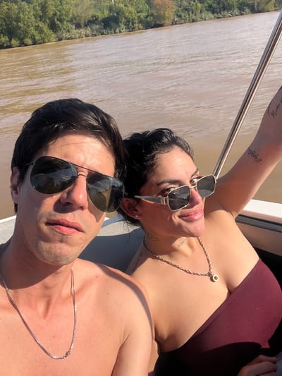 Two individuals, a man and a woman, are sunbathing on a boat in San Fernando, Argentina. The waterway beside the boat is muddy. Both are wearing sunglasses. The woman is wearing a dark red top and a gold chain, and has tattoos on her arms and chest. The man is shirtless and also wears a chain necklace.