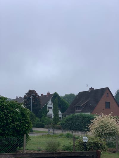 The image depicts a residential area in Rotenburg, Germany, on a hazy or overcast day. In the foreground, a chain-link fence runs horizontally across the bottom of the frame. Beyond the fence, a grassy area with some scattered plants and a lamp post is visible. A paved driveway or road is situated behind this area, with two ornamental metal gates providing access.
Several houses are visible in the background, characterized by their tiled roofs and brick or stucco exteriors. One prominent house on the right is made of red brick and has a pitched roof with a small window in the gable. To the left of this, other houses with varied architectural styles are partially obscured by lush greenery, including trees and bushes. Some of the houses are covered in ivy. The sky is a uniform, light gray, suggesting a cloudy or foggy atmosphere. No people are present in the scene.