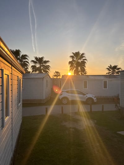 The image depicts an outdoor scene in Gavà, Spain, during either sunrise or sunset. The sun, low on the horizon and partially obscured by a palm tree, casts a strong golden light and visible lens flares across the frame. The sky transitions from a light blue at the top to a bright orange-yellow near the horizon. Two distinct, white contrails are visible in the upper left sky.

The scene features multiple mobile homes. On the left, the side of a light-colored mobile home with horizontal siding and several white-framed windows is prominent and brightly illuminated. Further back, several similar mobile homes are arranged, one with a small, light-colored fence or railing in front. A white compact SUV is parked on a paved pathway between the mobile homes in the midground, facing slightly left. Numerous palm trees, silhouetted against the bright sky, are scattered throughout the scene, rising above the structures. A patch of green grass is visible in the lower midground. No individuals are present.