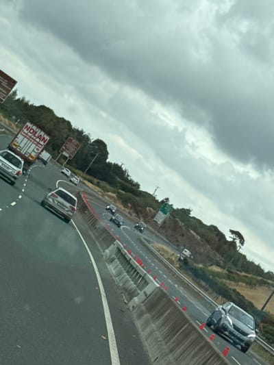A grey, overcast sky looms over a multi-lane highway in the New Ross Municipal District, Ireland. The highway curves to the right, with a concrete barrier on the right side of the road, adorned with red traffic cones. Several vehicles are visible on the highway, including a white truck with "NOLAN" written on its side, a white car, a silver station wagon, and a dark-colored SUV. The surrounding landscape features a wooded hillside on the right and a grassy embankment on the left. A street light is visible on the left side of the road. The image is taken from a moving vehicle, as indicated by the motion blur and the angle of the shot.