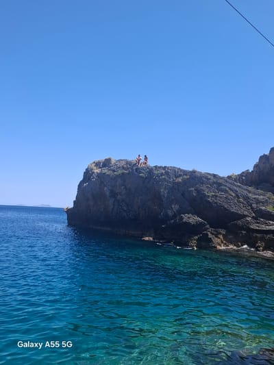 The image shows a rocky cliff jutting out into clear, blue water, likely the sea, under a clear blue sky. Two figures are perched on the edge of the cliff. The cliff face is dark and rugged. The water is translucent near the cliff, transitioning to a deeper blue further out. The scene suggests a coastal location, similar to cliffs found near Plakias, Greece.