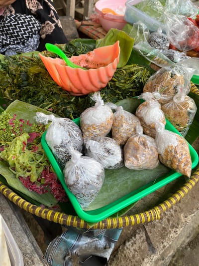 This image captures a close-up, overhead view of a market stall in Tambakreja, Indonesia. The scene is dominated by various food items arranged on banana leaves and in plastic bags. In the foreground, a green plastic tray holds several small, clear plastic bags filled with what appear to be seasoned grains or small fried items, some topped with a white powdery substance. To the left of the tray, there's a portion of a green banana leaf serving as a bed for thinly sliced green vegetables, scattered with small green seeds and pinkish flower petals. Behind this, a large pile of cooked green leafy vegetables is visible, with a bright orange, ribbed plastic bowl containing a small green utensil resting on top. In the background, more food items in plastic bags are visible, including some that appear to be dried or fried. The stall appears to be outdoors, with the suggestion of a woven basket base for the display and glimpses of a person's black and white patterned clothing at the top left. The overall impression is of a vibrant, local food market offering fresh produce and prepared snacks.
