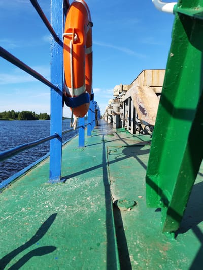 The image depicts a perspective shot from the deck of a ferry or boat, looking forward along its length. The foreground is dominated by the green, weathered deck, showing signs of wear and tear. To the left, a bright blue railing lines the edge of the deck, with a life preserver hanging from one of its blue supports. The life preserver is orange with white reflective stripes and a white rope. Beyond the railing, a calm, dark blue body of water stretches out, bordered by a lush green treeline on the distant shore.
The middle ground reveals the structure of the ferry, including industrial-looking metal components painted in various shades of gray and yellow, some showing rust. The perspective draws the eye towards the horizon, where the ferry's deck appears to extend indefinitely. A clear, bright blue sky with a few wispy white clouds is visible above.
In the far distance, a single person can be seen walking along the deck, providing a sense of scale. The strong shadows cast on the deck suggest it is a sunny day, likely late morning or early afternoon. The overall mood is one of transit and observation, with the harsh industrial elements of the ferry contrasted by the natural beauty of the water and landscape. The location is identified as Nova, Russia. No visible text is present in the image.
