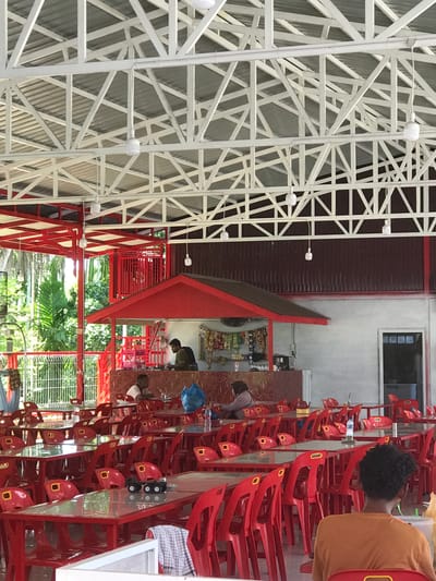 A casual, open-air restaurant scene in Bulo Gogo, Indonesia. The restaurant features a white, lattice-structured roof and red accents throughout. Rows of red plastic chairs are stacked at tables, and several individuals are seated, appearing to be patrons or staff. A bar area displays stocked shelves with food items. Outside, lush greenery is visible through the open walls of the restaurant.