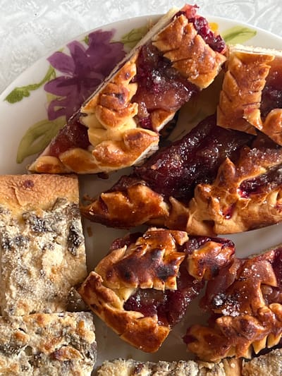The image shows a plate of baked goods. Some pieces are wedge-shaped, filled with a dark red jam, and topped with decorative pastry. Other pieces are rectangular and covered in a sugared topping with visible raisins. The plate is white with floral decorations, including a purple flower. The background is a neutral, textured surface. The location of Чернышово, Russia is not evident from the image.