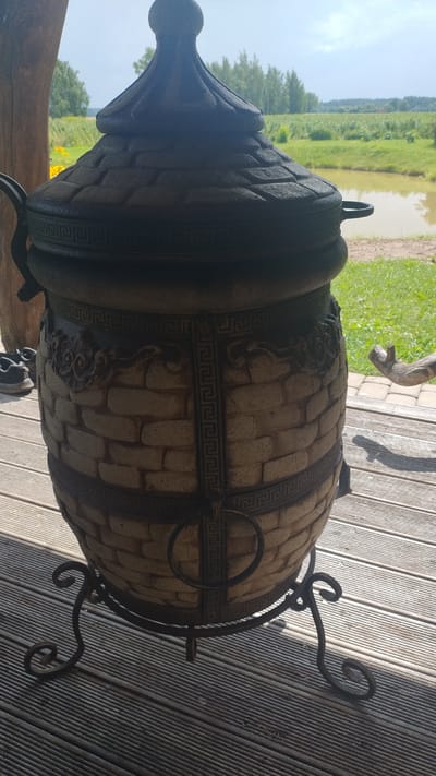 A tandoor oven stands on a wooden deck outdoors. The tandoor is made of tan brick with a dark lid and decorative metal bands. It is supported by a black metal stand with ornate legs. In the background, there is a body of water, green grass, trees, and a bright sky. The scene appears to be in Kalnrozes, Latvia, given the context of the request.