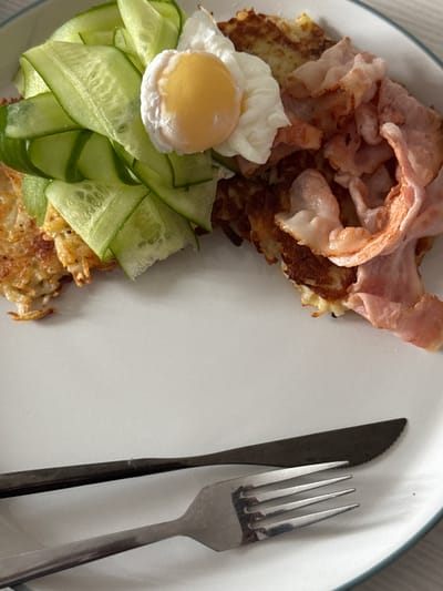The image shows a plate of food, likely breakfast, on a white plate with a blue rim. The food consists of potato pancakes, crispy bacon, sliced cucumber, and a poached egg. A knife and fork are placed on the plate, ready for eating. The background is a light-colored surface, possibly a table. There are no people visible in the image. The scene is domestic and appears to be well-lit. There is no information in the image to suggest any connection to the city of Osentsy, Russia.