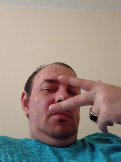 A man is shown in a low-angle, indoor selfie. He is wearing a turquoise shirt. His right hand is raised, with his index and middle fingers extended to form a peace sign gesture in front of his eye. He is wearing a ring with a black stone on his right ring finger. The background is a plain, light-colored wall.