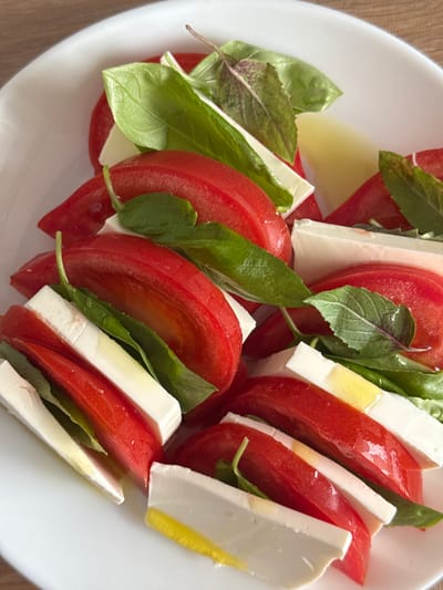 A plate of Caprese salad is presented on a wooden surface. The salad consists of sliced tomatoes, fresh mozzarella cheese, and basil leaves, arranged in alternating layers. The ingredients are glistening, suggesting they are dressed with olive oil and seasoning. The image is a close-up, focusing on the texture and vibrant colors of the food. There are no people or animals visible in the frame, and the setting is purely culinary. The location in Gamovo, Russia, is not visually represented in the image.