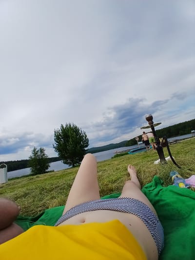 The image depicts a waterside scene, likely taken from a low angle, focusing on a person's legs and torso in the foreground. The individual is lying on a green blanket, wearing striped shorts and a yellow top. To the right of the frame, another person's bare feet are visible, also resting on a green blanket. Further back, in the middle ground, is a hookah, a body of water, a wooden pier, and several individuals standing on or near the pier. Two men are visible on the pier; one is wearing green shorts and appears to be interacting with the hookah. A third person is standing near the water. The background consists of a tree-lined shore and hills under a cloudy sky. The location is identified as Revda, Russia.