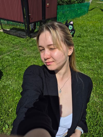 A young woman is captured in a selfie outdoors. She is wearing a black blazer over a white top and a silver necklace with a pendant. Her hair is blonde and pulled back, with a black hair clip shaped like a cat. She has a lip ring and a piercing in her ear. The background shows a green lawn, a red fence, and a glimpse of a building. The lighting suggests it is a sunny day. The image appears to be taken in the city of Sars, Russia, as indicated by the presence of a traditional wooden fence and a corrugated metal structure.