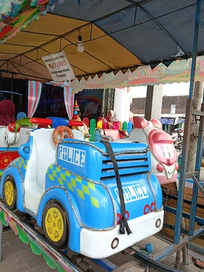 An outdoor scene in Sumberejo, Indonesia, depicts a children's amusement ride operating under a yellow and blue fabric canopy supported by a metal frame. The foreground features a blue and white miniature police car-themed ride cart, displaying "POLICE" in white block letters on its sides and rear, along with a green and yellow checkerboard pattern. Yellow wheels are visible. A black strap hangs from the rear of the police car cart. To the right, a white and pink ride cart designed like a cartoon face is visible.

A woman wearing a pink long-sleeved top and a light-colored hijab is seated in a ride cart to the left of the pink-faced cart, looking down at a mobile phone in her hands. Another person, largely obscured, is seated in a red-colored cart further to the left, wearing a maroon hijab. A white sign, hanging from the canopy's metal frame, reads "KERETA PANGGUNG Rp 7.000 DURASI MAKSIMAL 5 MENIT" (Stage Train Rp 7,000 Maximum Duration 5 Minutes). Another banner with colorful text "KIDS [partial text]" and "JAM 09-21.00" (9 AM - 9 PM) is visible on the right side of the canopy. The background shows a large white pillar, followed by residential or commercial buildings with dark roofs under an overcast sky. A blue shopfront sign is discernible in the far background.