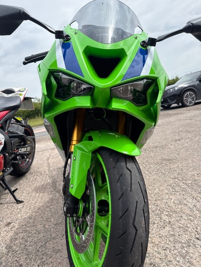 A close-up, low-angle shot captures the front of a bright green Kawasaki motorcycle, parked on a paved surface. The motorcycle features dual headlights, gold-colored front suspension forks, and a green front wheel with visible brake disc and calipers. A portion of a black car is visible in the background to the right, and another motorcycle, red and white with "CBR RR" branding, is partially visible to the left. The environment appears to be outdoors, with a cloudy sky overhead and a weathered asphalt surface beneath the vehicles. There is no indication of a specific location within Much Dewchurch, United Kingdom.
