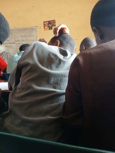 An indoor scene in Ugi Majowel, Nigeria, depicts multiple individuals seated in what appears to be a classroom. The camera perspective is from behind the seated individuals. In the immediate foreground, the back of a dark green chair or desk is visible. To the right, the back and right shoulder of a person wearing a brown garment with short dark hair are present. Centrally, a person with short dark hair is seated, facing forward, wearing a light gray or olive-colored garment. A hand with lighter skin tone is touching the top of this person's head. Further to the left and behind, another person wearing a light blue or green garment is partially visible, along with the top of a head wearing dark, possibly knitted, headwear and a person in a red shirt. The background features a light yellow wall with a white board. The white board displays handwritten mathematical equations, including "L = 30 + (1-t)LxK". Two small posters with colored illustrations are affixed to the wall above and to the right of the whiteboard. The lighting is diffused, indicating an interior setting.