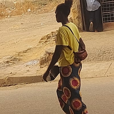 The image captures an outdoor scene during what appears to be bright daylight, suggested by the strong illumination and shadows. The primary subject is a person, likely dark-skinned, positioned in a bent or squatting posture, facing downwards and slightly to the left of the frame.
This individual is wearing a bright yellow, possibly sleeveless, top or tunic, with a dark strap visible across their back, possibly from a bag. Their head is covered with a dark fabric or hairstyle, and their hair appears dark.
To the right of this person, a section of a vibrant, dark-colored fabric is visible, patterned with repeating circular motifs in red and gold, resembling stylized suns or flowers. This could be a piece of clothing worn by the person (e.g., a skirt or wrap) or another person standing nearby.
The ground and background surfaces are predominantly light brown or tan, appearing rough and textured, consistent with a dusty street, unpaved area, or an old wall. In the upper left corner of the image, there's a dark rectangular window with a light-colored security grill of horizontal and vertical bars. Below this window, a white cloth or garment is draped, and a small, light-colored object (possibly a shoe or another piece of clothing) rests on the ground.
The overall impression is of an everyday moment, possibly in a warm climate, with the person engaged in an unspecified activity or simply resting. No text is visible in the image.