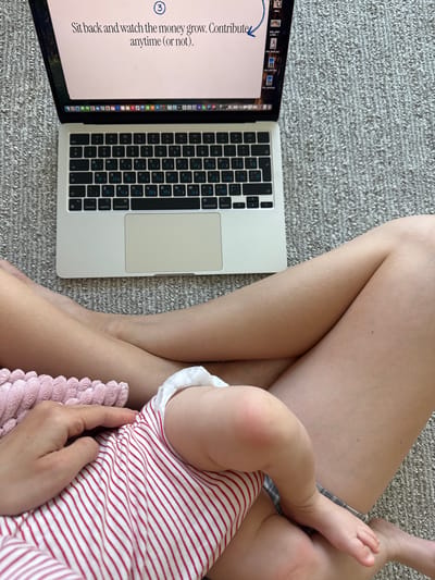 The image shows a person sitting on a gray carpet with a laptop open on their left, displaying text about money growth. The person is holding a baby dressed in a red and white striped outfit. The baby's leg and foot are visible. The scene appears to be indoors. There is no indication of the location being Springfield, United States of America.